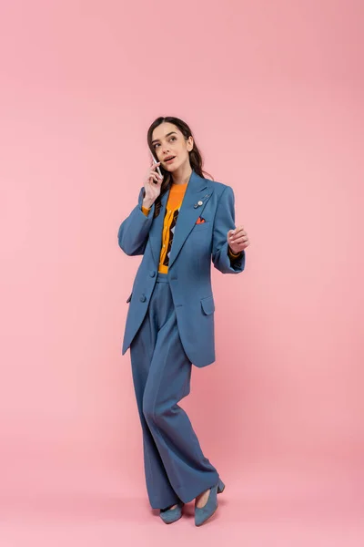 Full length of young brunette woman in blue pantsuit talking on smartphone on pink — Stock Photo