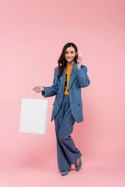 Full length of smiling woman in blue pantsuit talking on smartphone and holding shopping bag on pink — Stock Photo
