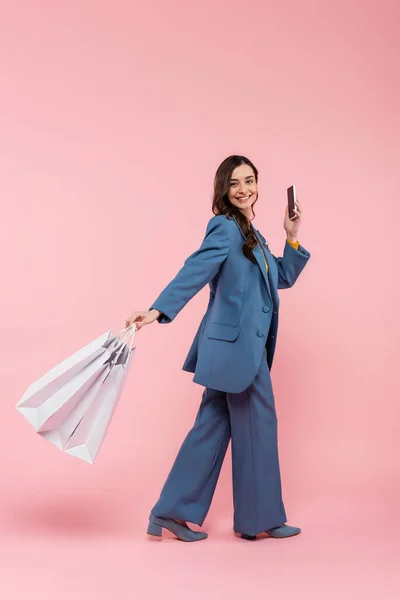 Full length of smiling woman in blue pantsuit holding smartphone and shopping bags on pink — Stock Photo