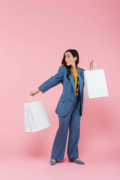 Full length of emotional young woman in blue pantsuit holding shopping bags on pink — Stock Photo