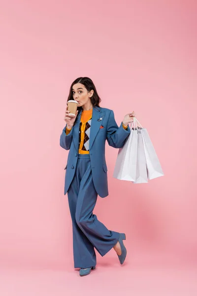 Full length of young woman in blue pantsuit holding shopping bags and drinking coffee to go on pink — Stock Photo