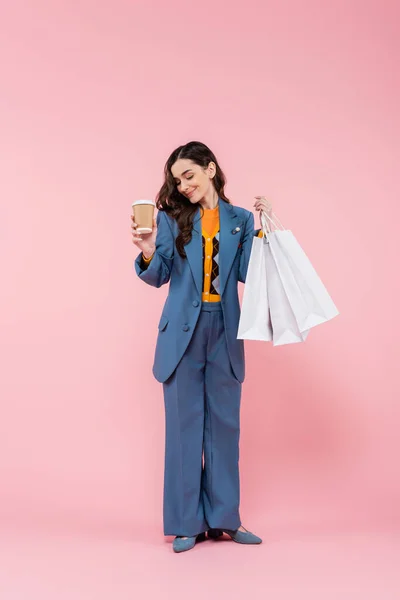 Full length of pleased young woman in blue pantsuit holding shopping bags and coffee to go on pink — Stock Photo