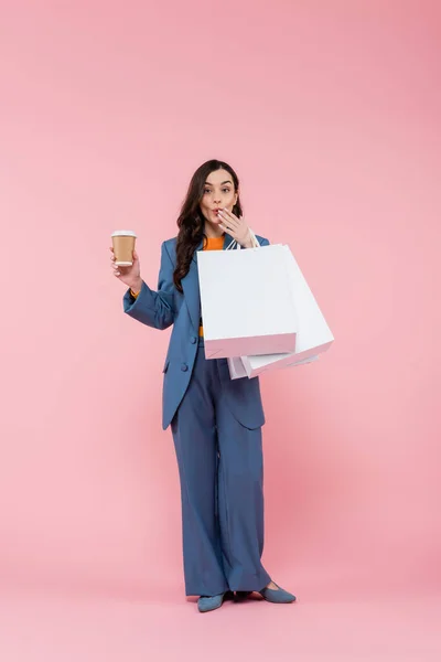 Full length of emotional woman in blue pantsuit holding shopping bags and coffee to go while covering mouth on pink — Stock Photo