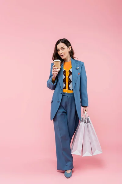 Full length of brunette young woman in blue pantsuit holding shopping bags and coffee to go on pink — Fotografia de Stock