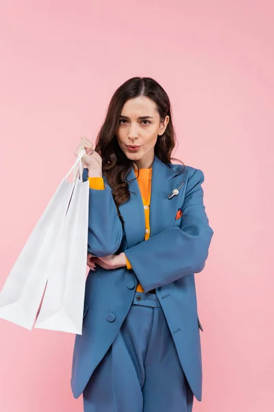 Young woman in blue blazer holding shopping bags and pouting lips isolated on pink — Stock Photo