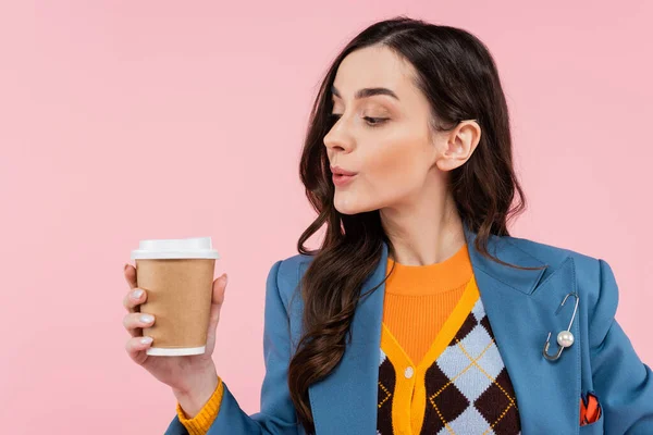 Молодая женщина в синем блейзере смотрит на бумажную чашку с кофе, чтобы пойти изолированы на розовый — стоковое фото