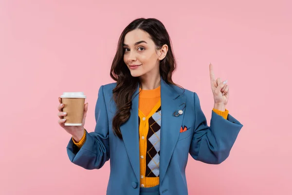 Young woman in blue blazer holding paper cup with coffee to go and pointing with finger isolated on pink — Stock Photo