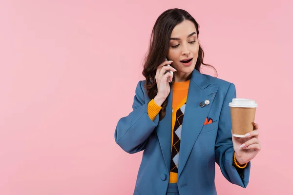 Amazed young woman in blue blazer talking on smartphone and looking at paper cup with coffee to go isolated on pink — Fotografia de Stock