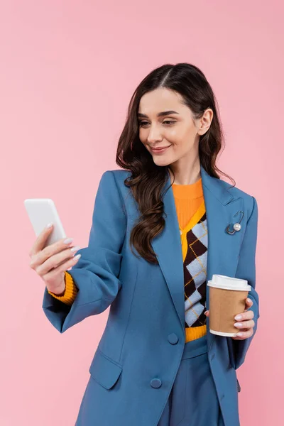 Cheerful woman in blue blazer holding smartphone and paper cup isolated on pink — Fotografia de Stock