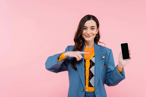 Cheerful young woman in blue blazer pointing at smartphone with blank screen isolated on pink — Stock Photo