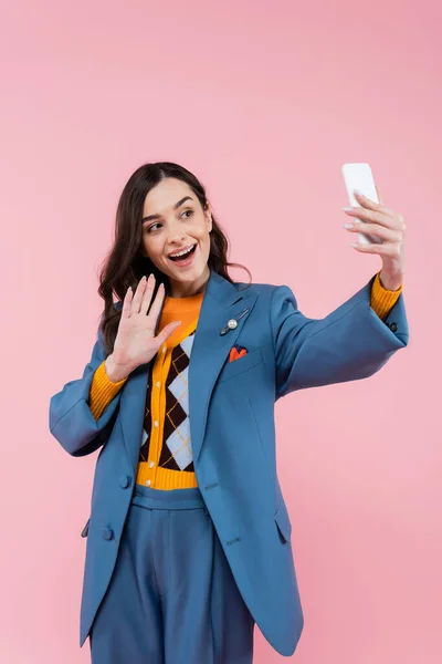 Glückliche junge Frau im blauen Blazer winkt bei Videoanruf isoliert auf rosa — Stockfoto