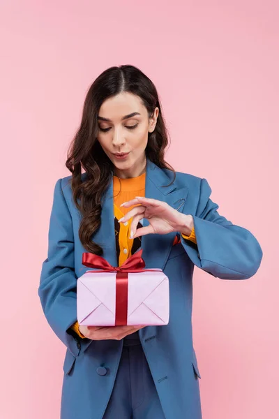 Mujer joven en blazer azul mirando regalo envuelto aislado en rosa - foto de stock