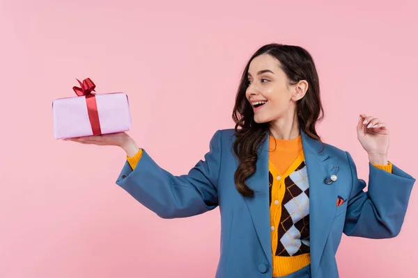 Sonriente joven en blazer azul mirando regalo envuelto aislado en rosa - foto de stock