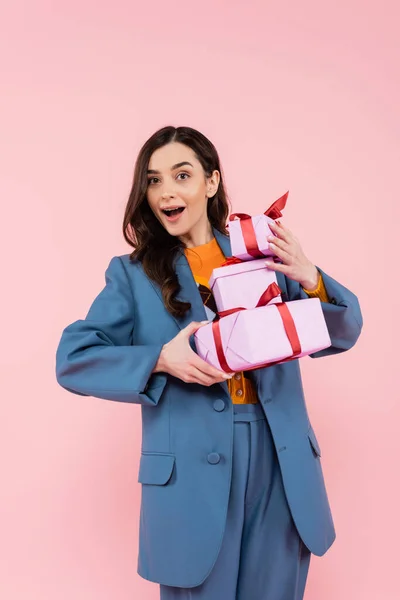 Mujer impresionada en chaqueta azul sosteniendo cajas de regalo mientras mira la cámara aislada en rosa - foto de stock