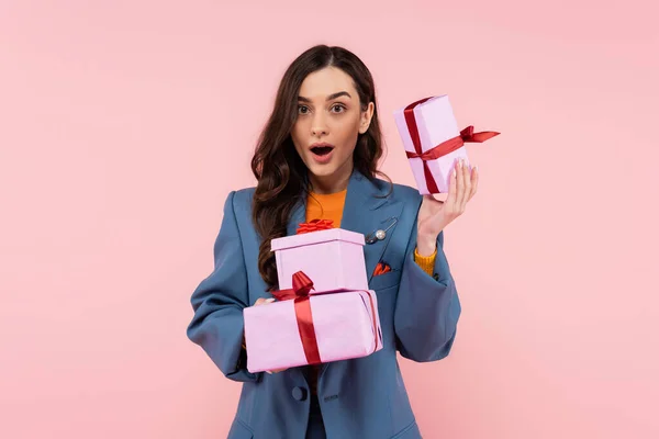 Amazed woman in blue stylish blazer holding presents while looking at camera isolated on pink — Stock Photo