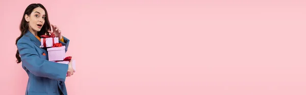 Astonished woman in blue blazer holding gift boxes and looking at camera isolated on pink, banner — Stock Photo