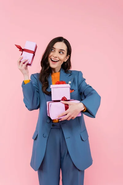 Mujer sorprendida y feliz en traje azul sosteniendo regalos mientras está de pie con la boca abierta aislada en rosa - foto de stock