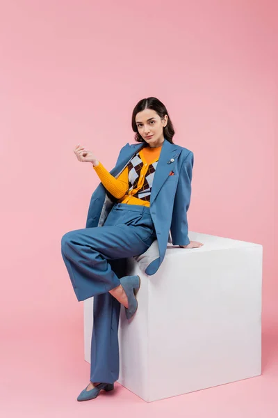 Full length of trendy brunette woman in blue suit posing near white cube and looking at camera on pink background — Stock Photo