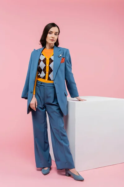 Full length of brunette woman in trendy casual attire standing near white cube on pink background — Stock Photo