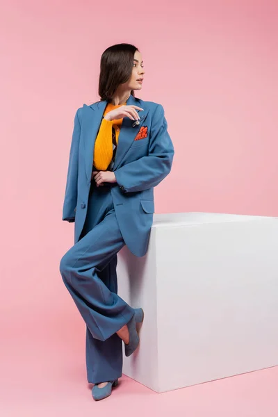 Full length of brunette woman in blue suit looking away while standing near white cube on pink background - foto de stock