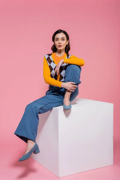 Full length of woman in blue pants and trendy cardigan sitting on white cube and looking at camera on pink background — Stock Photo