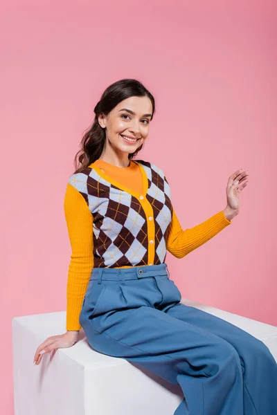 Cheerful woman in blue pants and orange cardigan sitting on white cube and waving hand isolated on pink — Foto stock