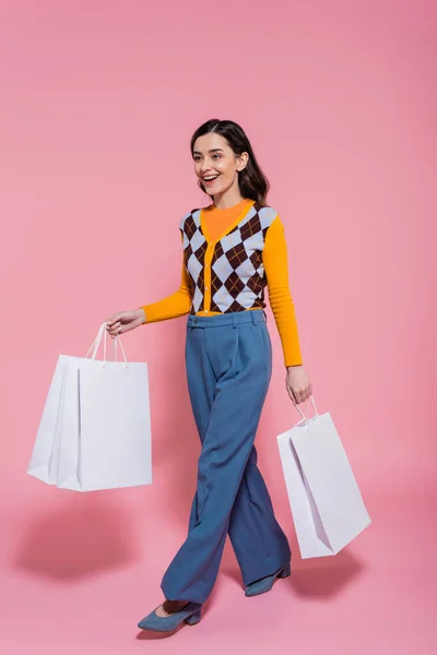 Full length of impressed woman in trendy cardigan and blue pants walking with shopping bags on pink background — Stock Photo