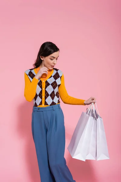 Impressed woman in fashionable cardigan and blue pants looking at white shopping bags on pink background — Fotografia de Stock