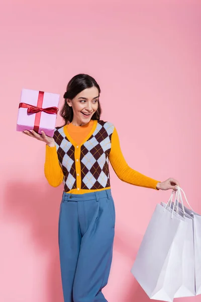 Mujer alegre en cárdigan de moda y pantalones azules sosteniendo bolsas de compras y caja de regalo mientras mira hacia otro lado sobre fondo rosa - foto de stock