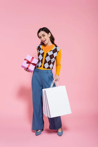 Piena lunghezza di donna soddisfatta in abito alla moda in piedi con scatola regalo e borse della spesa su sfondo rosa — Foto stock