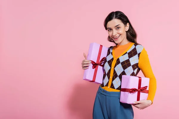 Young woman in bright cardigan holding gift boxes and smiling at camera on pink background — Stock Photo