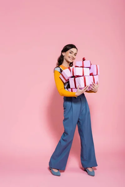 Full length of cheerful woman in blue pants holding plenty of presents and looking at camera on pink background — Stock Photo