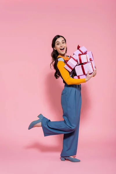 Full length of amazed and happy woman in blue pants holding pile of gifts on pink background — Stock Photo