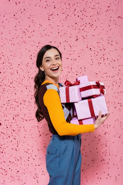 Cheerful woman in stylish clothes holding gift boxes while looking at camera on pink background — Stock Photo