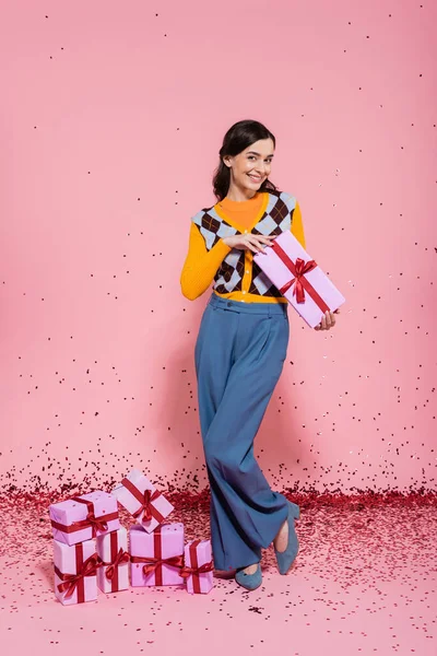 Full length of pleased woman posing with present near festive confetti and gift boxes on pink background — Foto stock