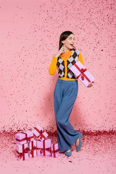Full length of surprised woman in trendy outfit standing with present near gift boxes and festive confetti on pink background — Stock Photo