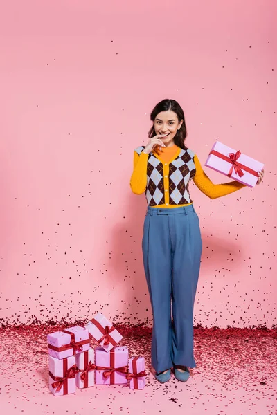 Pleased woman in blue pants holding present and looking at camera near confetti and gift boxes on pink background — Stock Photo