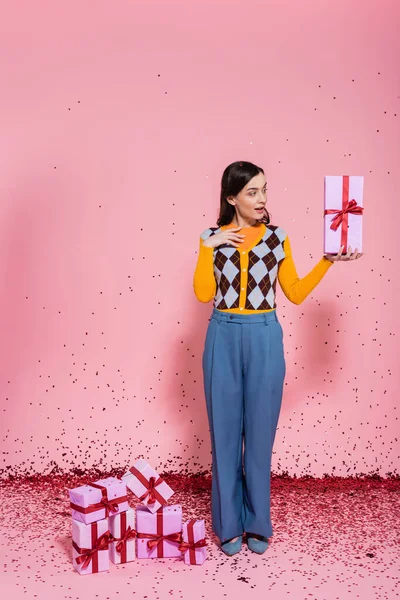 Femme surprise en pantalon bleu et cardigan tendance regardant présent près de confettis festifs et boîtes-cadeaux sur fond rose — Photo de stock