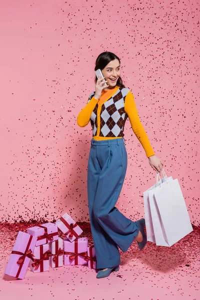 Femme souriante et branchée avec des sacs à provisions parlant sur smartphone près de boîtes-cadeaux et confettis sur fond rose — Photo de stock