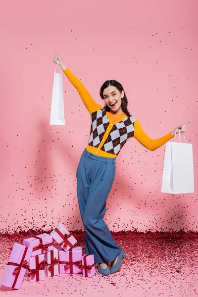 Cheerful and stylish woman holding white shopping bags near gift boxes and festive confetti on pink background — Stock Photo