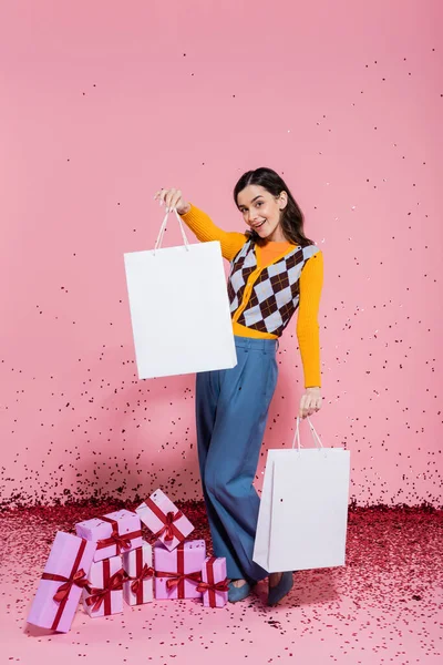 Full length of fashionable woman posing with white shopping bags near gift boxes and festive confetti on pink background — Stock Photo