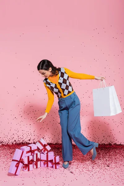 Full length of fashionable woman with shopping bags looking at gift boxes near festive confetti on pink background — Stock Photo