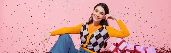 Cheerful woman in fashionable attire posing near gift boxes and festive confetti on pink background, banner — Stock Photo
