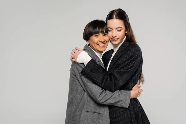 Middle aged asian woman in stylish clothes embracing brunette daughter while looking at camera isolated on grey — Stock Photo