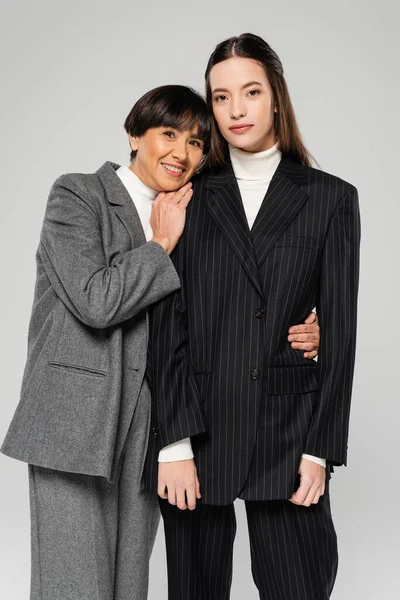 Happy asian woman in grey suit hugging young daughter while looking at camera isolated on grey — Stock Photo
