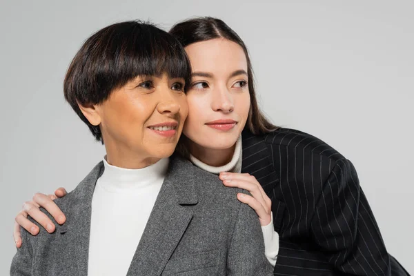Positive asian mother and daughter in white turtlenecks and blazers looking away isolated on grey — Fotografia de Stock