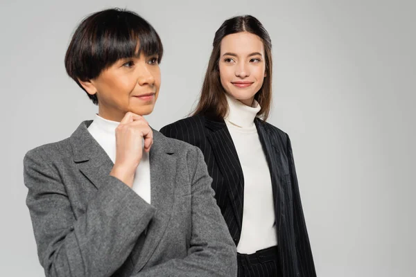 Positive woman looking at thoughtful asian mother holding hand near chin and looking away isolated on grey — Stock Photo