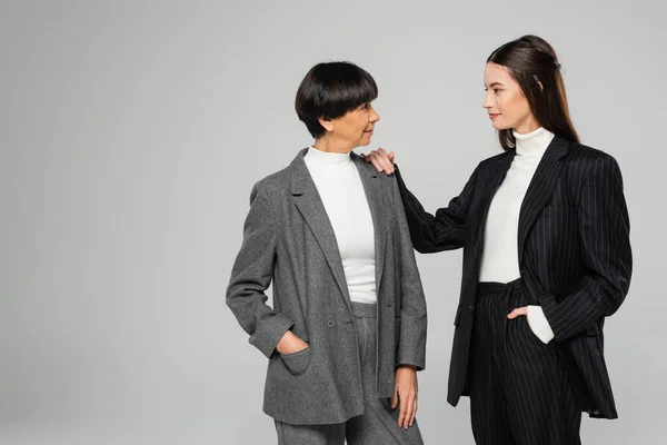 Brunette woman in black suit touching shoulder of elegant asian mother isolated on grey — Stock Photo