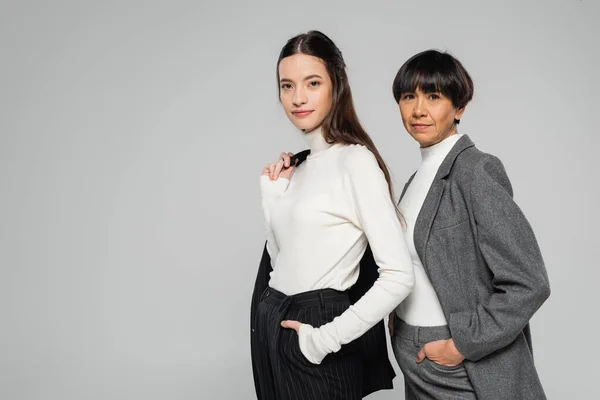 Positive asian mother and daughter in formal wear posing with hands in pockets while looking at camera isolated on grey — Photo de stock