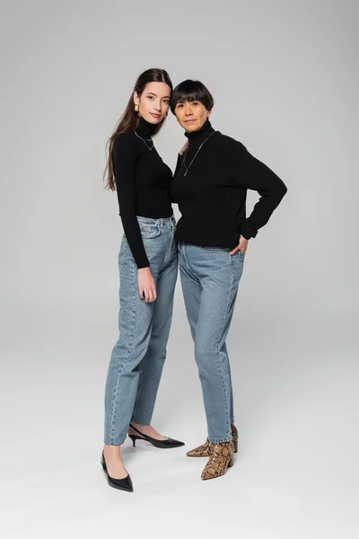 Full length of trendy asian mother and daughter in black turtlenecks and jeans posing on grey background — Stock Photo